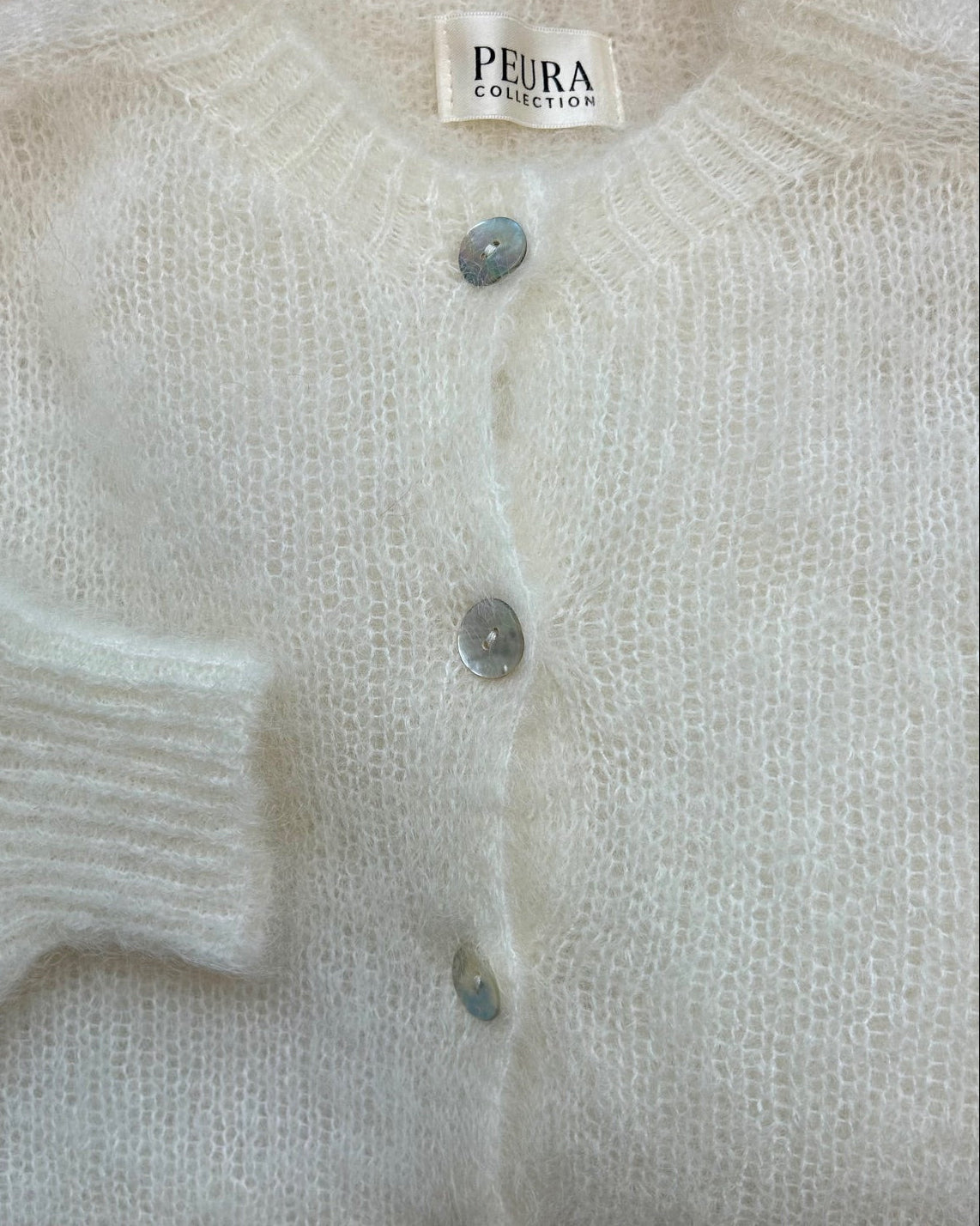 Peura Collection Nelly luonnonvalkoinen mohairneuletakin yksityiskohdat. Pehmeä, pörröinen neule, jossa on pyöreä päätie, helmiäisnapit ja pitkät hihat.